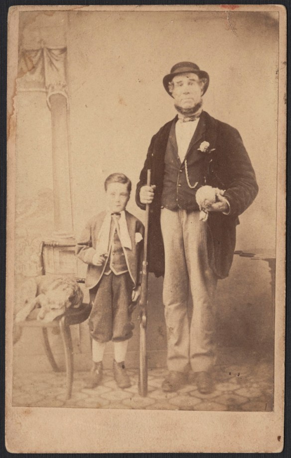 A black-and-white photographic portrait of a dog resting on a chair next to a boy and a man holding a rifle. 