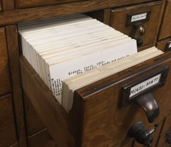 A brown card catalogue with one drawer open. The drawer contains several catalogue cards. A catalogue card with an author’s name and date of birth printed at the top is visible. It reads Bishun, Cyril, 1922-.