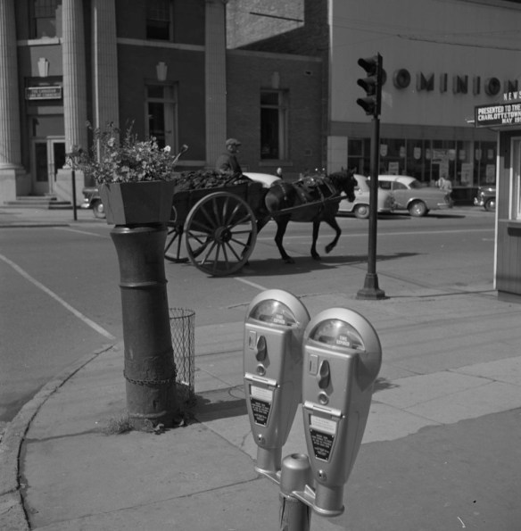 A black-and-white photograph of a double-headed parking meter on a street. In the background there are parked cars and a passing horse-drawn cart. 