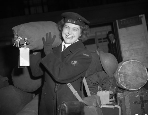 A black-and-white photograph of a smiling member of the Women’s Royal Canadian Naval Service carrying a large bag on her shoulder. 