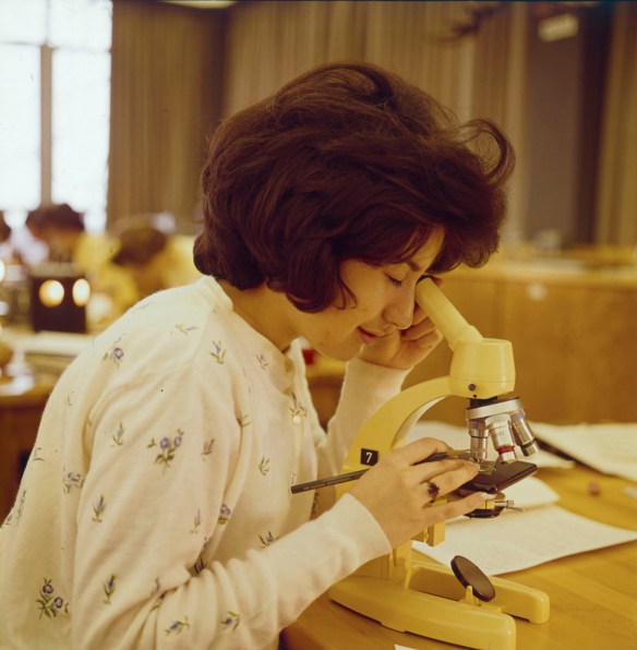 A colour photograph of a woman in a university classroom looking through a microscope. 