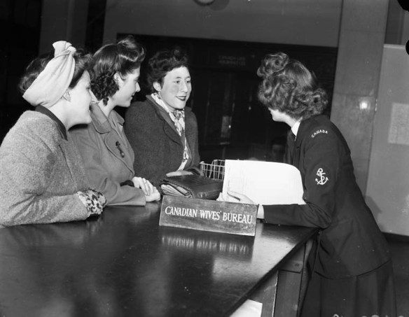 A black-and-white photograph of a woman in a military uniform leaning across a counter to interview three women beside a sign that reads “Canadian Wives’ Bureau.” 
