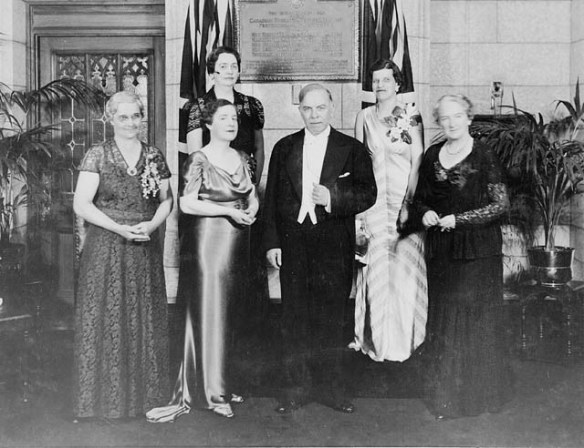 Five women in gowns wearing corsages and one man in a tuxedo standing in front of a plaque.