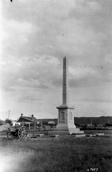 A black-and-white photograph of an obelisk monument with a wheeled cart nearby.