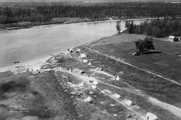 A black-and-white photograph of an aerial view of a riverbank with narrow roads, tents and people walking. On the right are trees and a barn-like building. 
