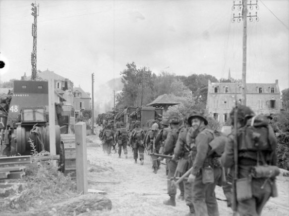A black-and-white photograph showing a column of soldiers marching up a street in a damaged village.