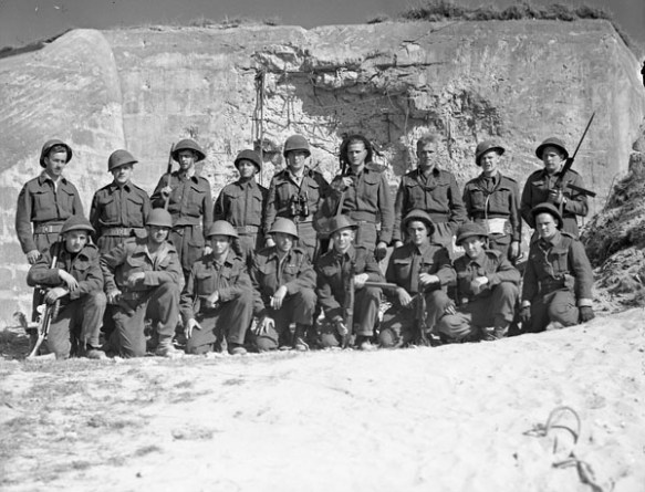 A black-and-white photograph showing two rows of sailors in battle dress, front row crouching, with a damaged fortified concrete structure behind them.