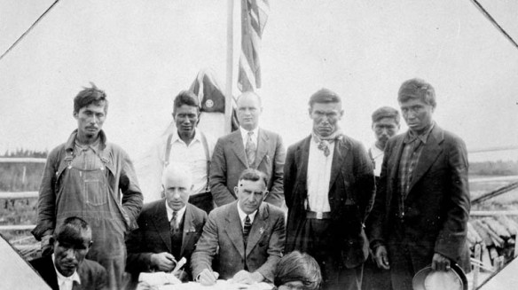 A black-and-white photograph of First Nations men and government officials posing in front of treaty documents, with a flag in the background.