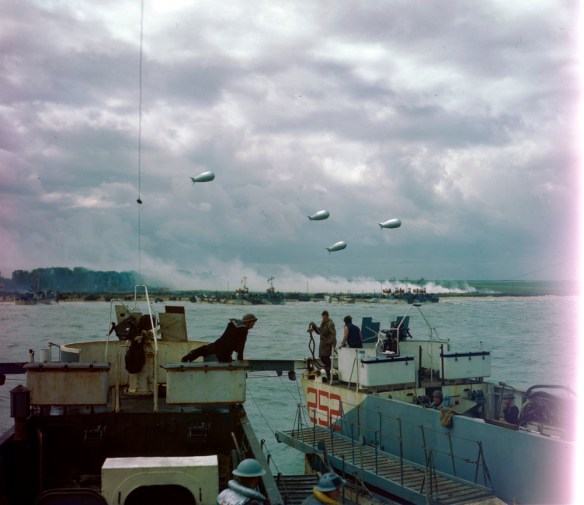 A colour photograph showing a landing craft approaching the beach, with smoke coming from a village and barrage balloons overhead.