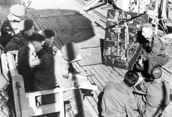 A black-and-white photograph of an officer briefing a small group of non-commissioned officers with a map of a village.