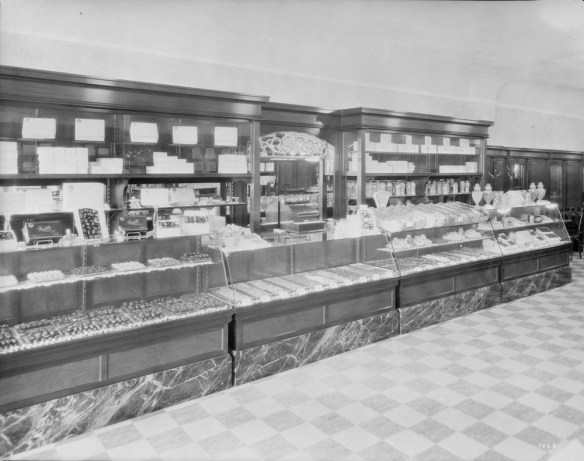 Four glass cases packed with baked goods form a long counter. Tall wooden shelves with a mirror in the middle are lined with boxes. The floor has a checkerboard pattern.