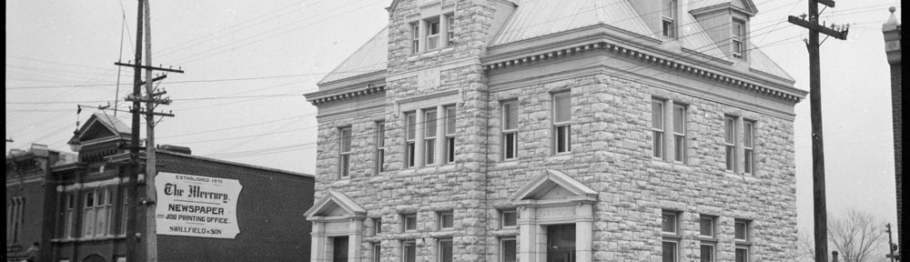A black-and-white photograph of a large stone building. In front of the building, there are men walking on the sidewalk. The sign on the building next door reads “The Mercury Newspaper.”