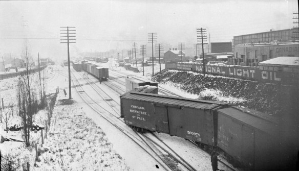 A black-and-white photograph of a partially derailed train in a train yard. Snow covers the ground and a city can be seen in the background.