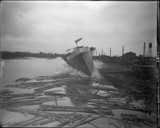 A black-and-white photo of the launching of a ship into the water, surrounded by logs and other debris.