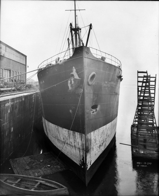 A black-and-white photo of the front of a large ship. There is a dent on the side of the hull that is furthest from the dock.