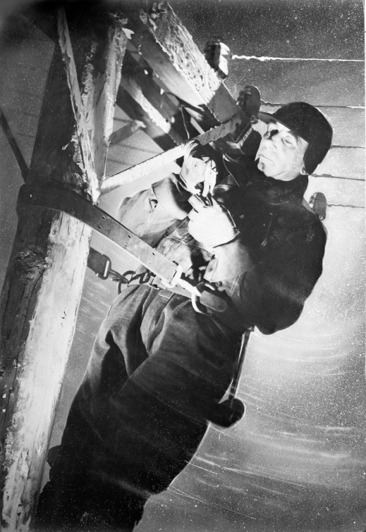A black-and-white photo of a man standing on a telephone pole and working on the wires. It is snowing, and he is wearing a winter coat, gloves and hat.