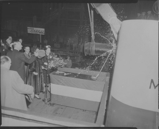 A black-and-white photo of men and women watching a champagne bottle being broken against the hull of a large ship. A man is holding his hat over the face of a woman wearing a fur coat.