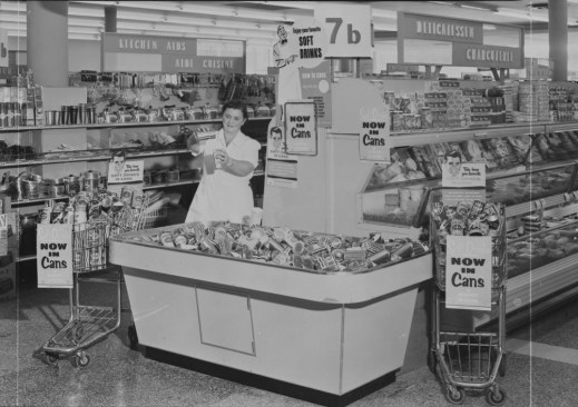 A black-and-white photo of a woman in a grocery story wearing a white dress and pouring a liquid from a can into a glass. She is standing behind a bin of canned drinks and two shopping carts with signs that read “Now in Cans.”