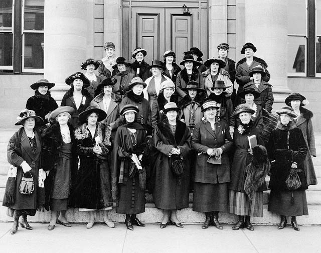 Black-and white photo of a group of women wearing hats and coats standing on the steps of a building.