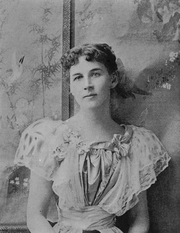 Black-and-white photo of a woman in a short-sleeved gown leaning against a wall.
