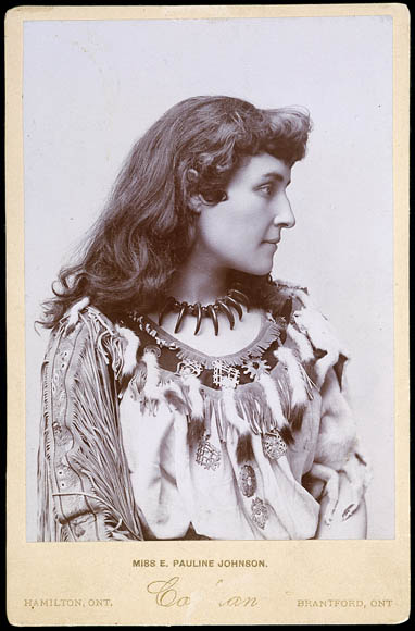 Black-and-white photo of a woman in profile. She is wearing a long-sleeved dress trimmed with tassels and fur along the sleeves.