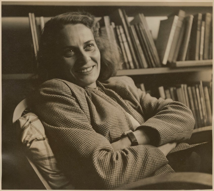 Sepia coloured photo of a smiling woman wearing a blazer. She is leaning back in a chair with her arms crossed.