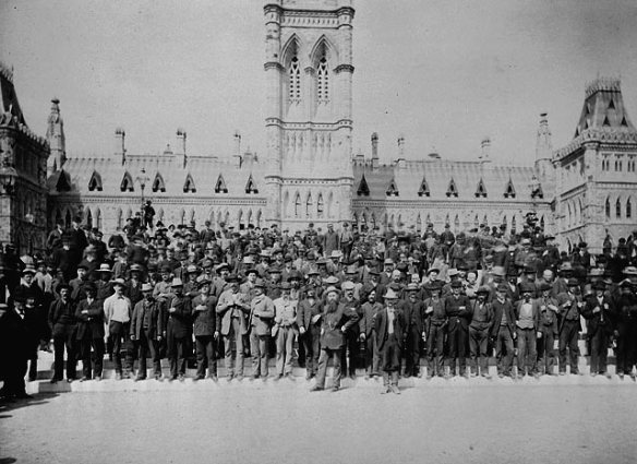 A black-and-white photograph of a large group of men standing in front of the Parliament buildings.