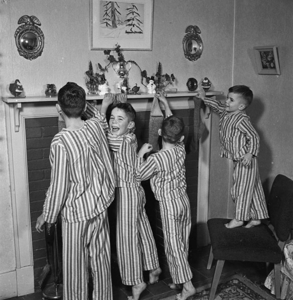 Photograph of four boys in matching striped pyjamas hanging Christmas stockings on a fireplace mantle.