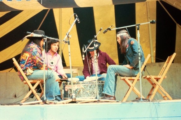Colour photograph of four men sitting on wooden chairs surrounded by microphones and facing each other, singing and drumming.