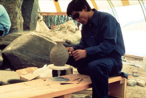 Colour photograph of a man in dark blue clothing wearing sunglasses and sitting on a wooden bench carving a vase. 
