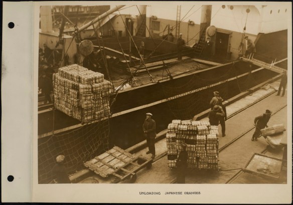 Port workers unloading crates of oranges from a ship, using cranes and carts.