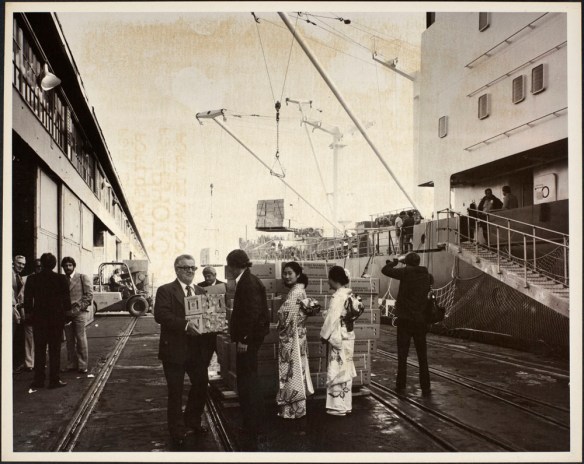 Photograph showing port officials holding boxes of mandarin oranges, two Japanese women in traditional dress standing by a pallet of orange boxes, and other people and equipment on a pier adjacent to a ship.