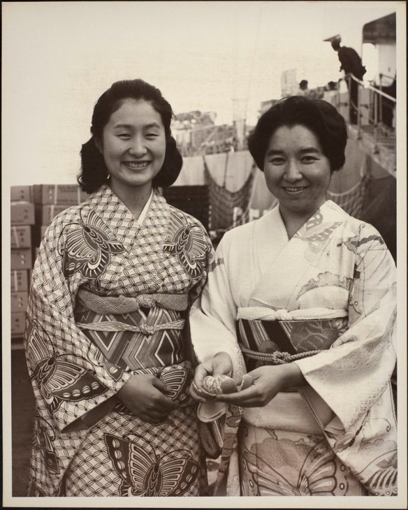 Photograph of two Japanese women wearing traditional dress, one of whom is holding a peeled mandarin orange. Crates of oranges and a ship are visible in the background.