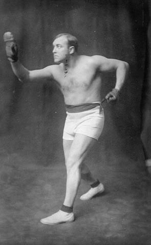 Black-and-white photo of a man wearing boxing gloves, shorts and shoes.