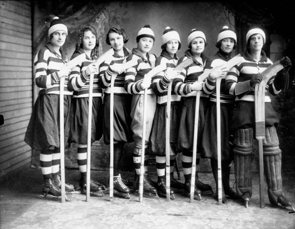 A black and white photo of a women’s hockey team lined up with the butt end of their sticks on the ground and dressed in their team uniform.