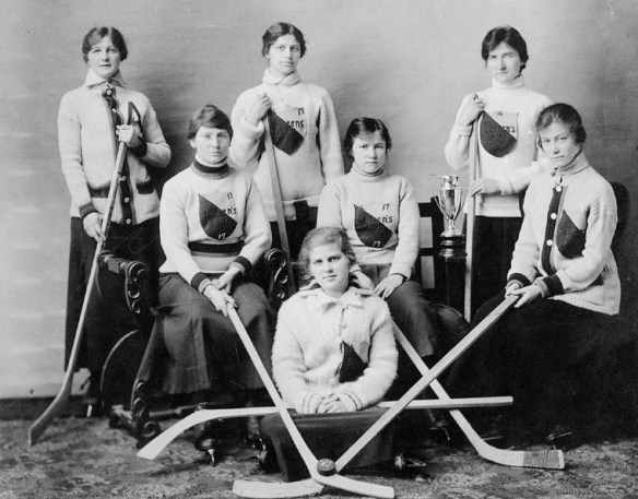 A black and white photo of a women’s hockey team. The women have team sweaters on and are holding their hockey sticks.