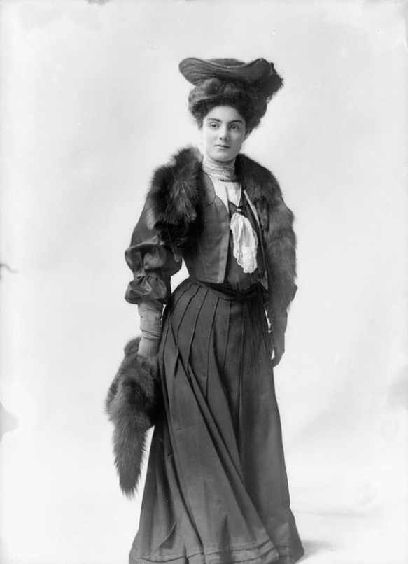 Black-and-white photo of a woman standing and facing the camera, wearing a dark-coloured dress, a fancy hat and a fur stole.