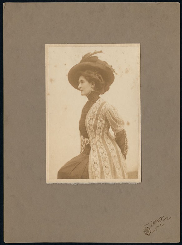 Side profile of Madge Macbeth in a black dress and white lace jacket wearing a feathered hat. 