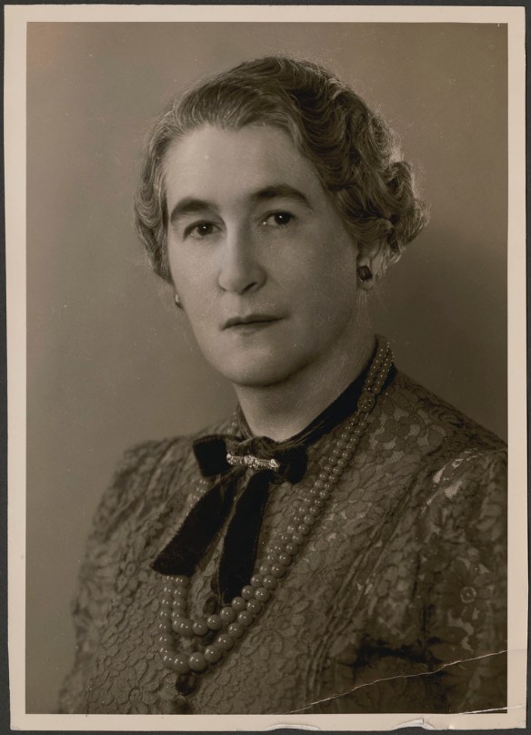 Portrait-style photograph of a woman wearing a lace blouse, jade beads and a diamond pin facing the camera. 