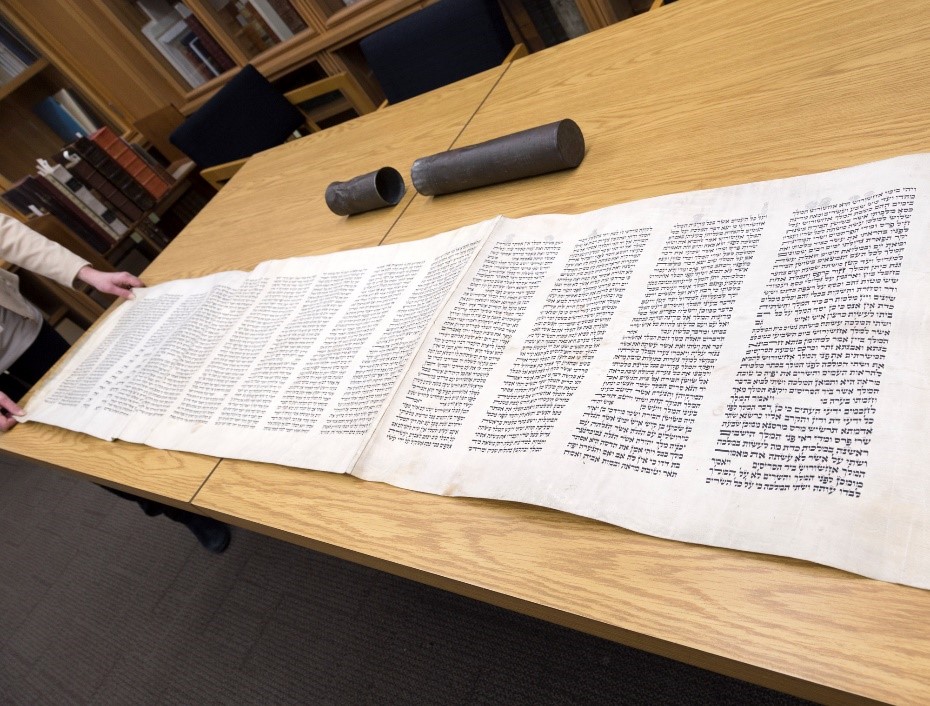 A colour photograph of a handwritten Hebrew manuscript on parchment completely rolled out across a table, approximately seven feet in length. Also on the table is the protective metal casing that originally housed the manuscript.