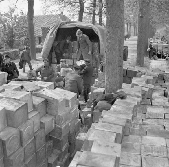 Several people unload food crates from the back of a military truck. Many crates are stacked in the foreground.