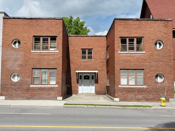 A colour photograph of a brown brick low-rise apartment building, with a white door. 