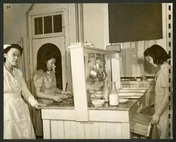Three women, one of whom is a nurse, are standing around a kitchen island on which there are trays, dishes and bottles of milk. Utensils are hanging from the rack that runs down the middle of the unit. 