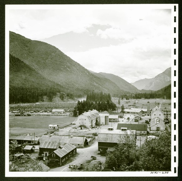 View of a small town surrounded by mountains. In the foreground are multiple buildings, and in the background on the left are rows of smaller houses.