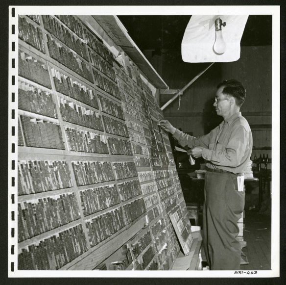 A man is standing in front of a large, tilted shelving unit filled with Japanese characters used in a printing press.