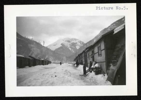 From right to left: Children in front of shiplap shacks with snow shovelled against walls. Internee in distance walking down makeshift “street.” Tar paper, to protect the shacks from the elements, is visible on the shack walls.