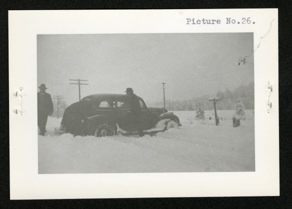 Three men and a car in a snowstorm: (from left to right) one man standing at the rear of the car, a second man bent over the back right tire, and a third man going towards the car to assist.