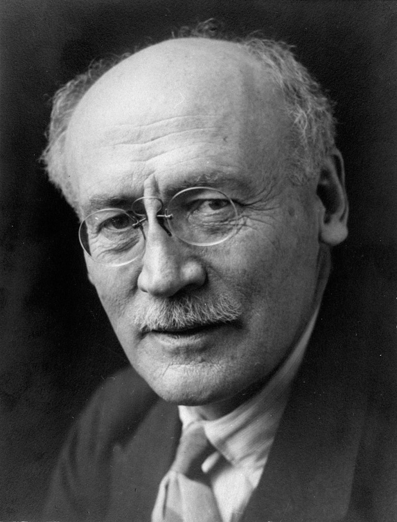 Black-and-white photo of a man’s face in close-up. He has a moustache and is wearing small round glasses.