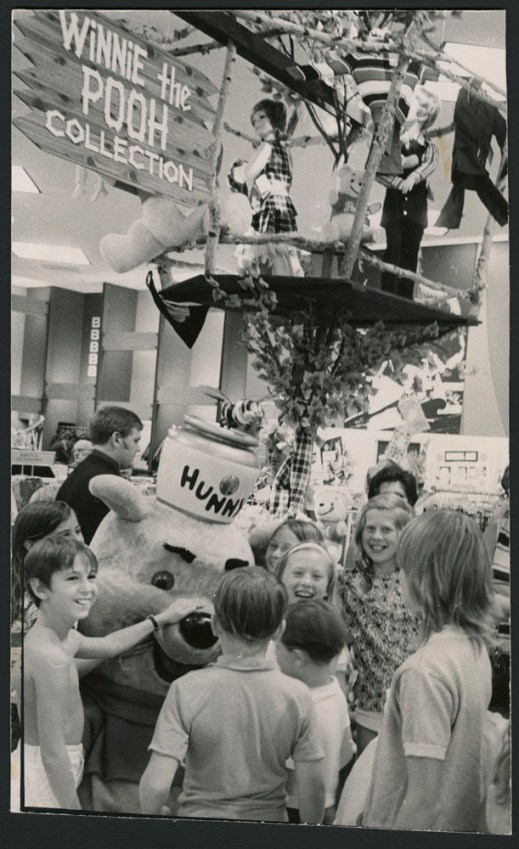 Black and white photograph showing children gathered around a large Winnie-the-Pooh mascot, with a treehouse and a “Winnie-the-Pooh Collection” sign in the background.