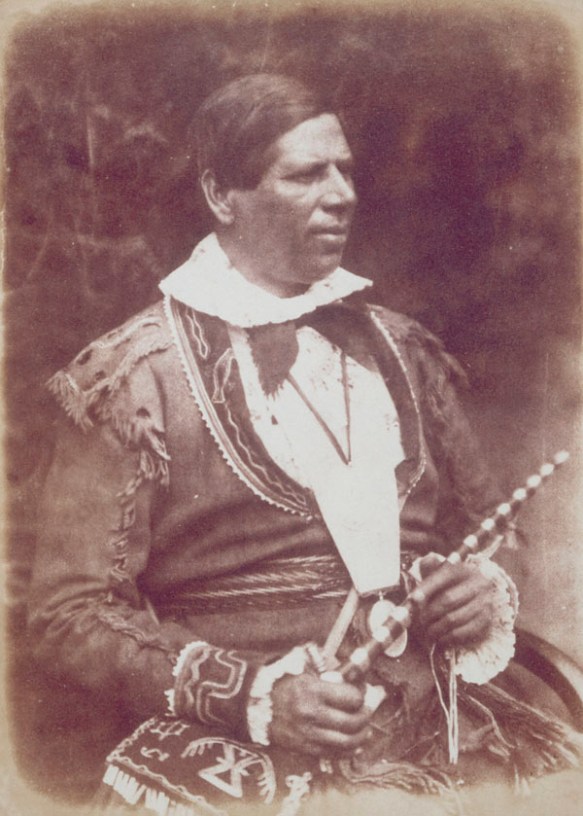 A sepia photograph of a seated man dressed in traditional quill design buckskin jacket and bag, holding a ceremonial pipe and a string of wampum beads. 
